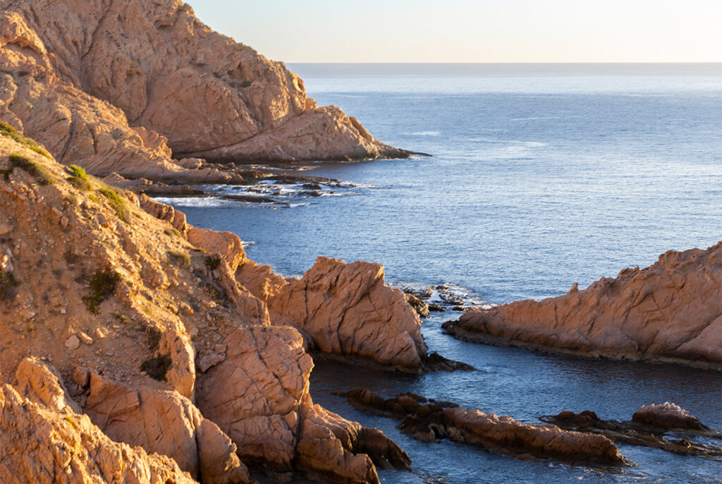 Sunlit rugged rocky coastline with jagged cliffs extending into calm blue ocean waters under a clear sky.